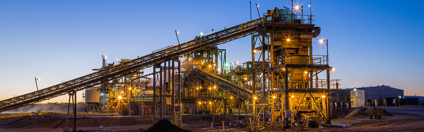 Mineral processing facility at dusk with conveyor belts, silos, and industrial lighting under a clear blue sky at dusk